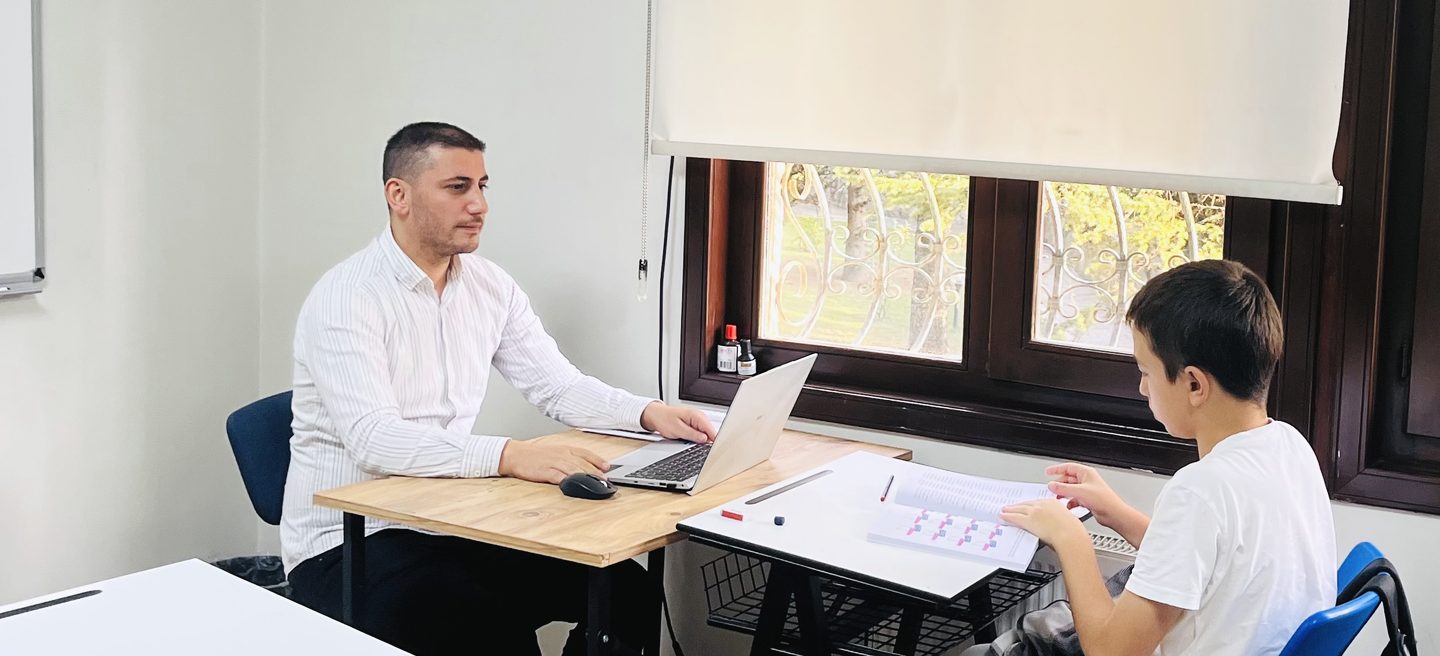 Sınıfta, eğitmen Hüseyin Taşpınar masasında dizüstü bilgisayarının başında oturuyor; pencerenin yanındaki masada bir öğrenci açık bir kitapçığı inceliyor.